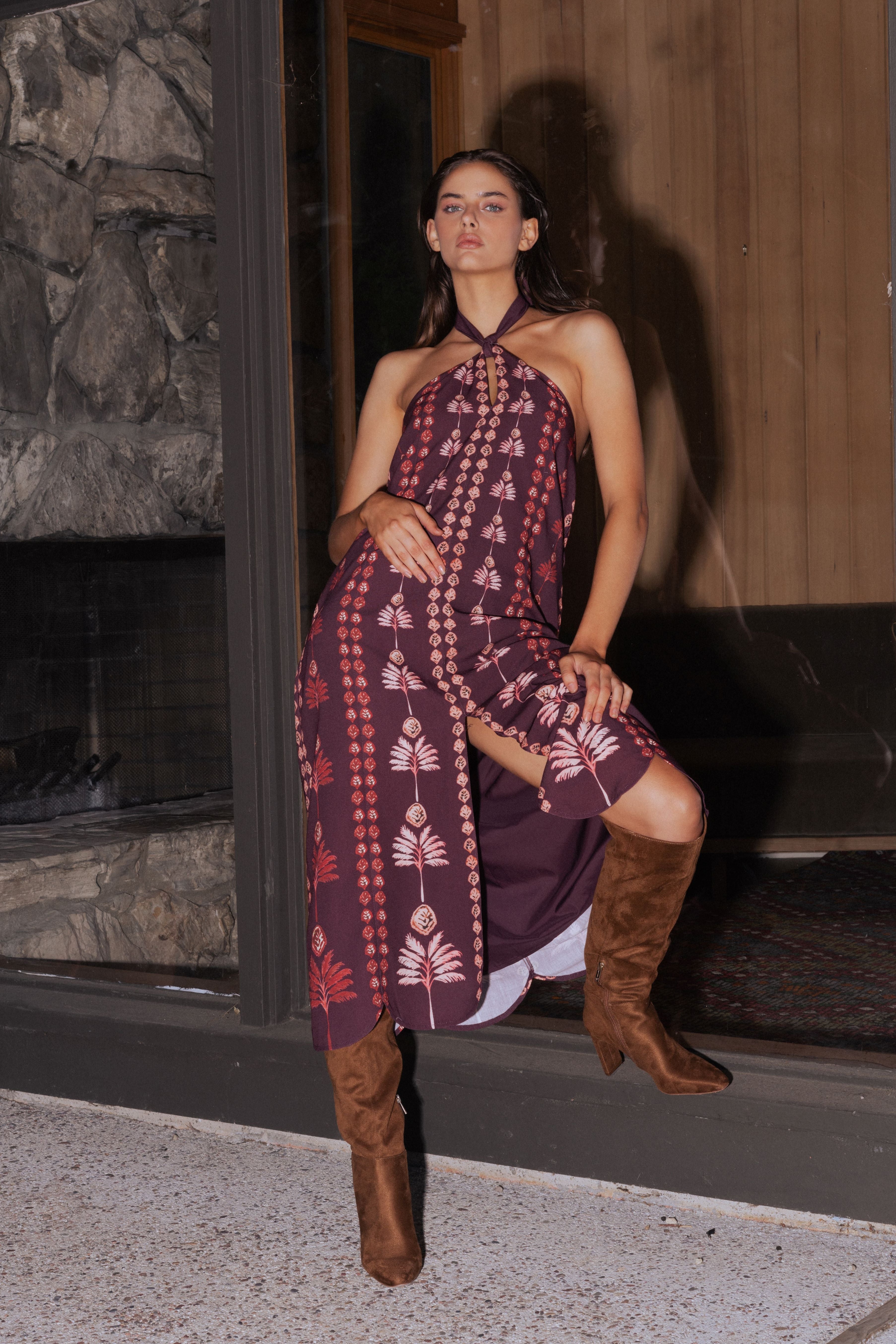 A woman models a halter-neck maxi dress with a deep purple base and a pink bohemian-style geometric pattern, paired with brown leather cowboy boots, posed in front of a rustic wooden and stone fireplace.