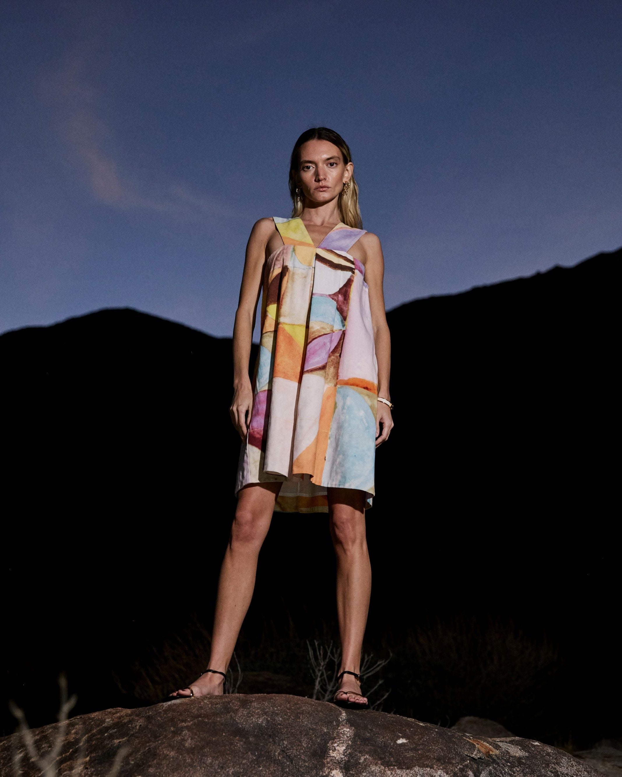Woman in a colorful dress standing on a rocky outcrop with mountains in the background at night.