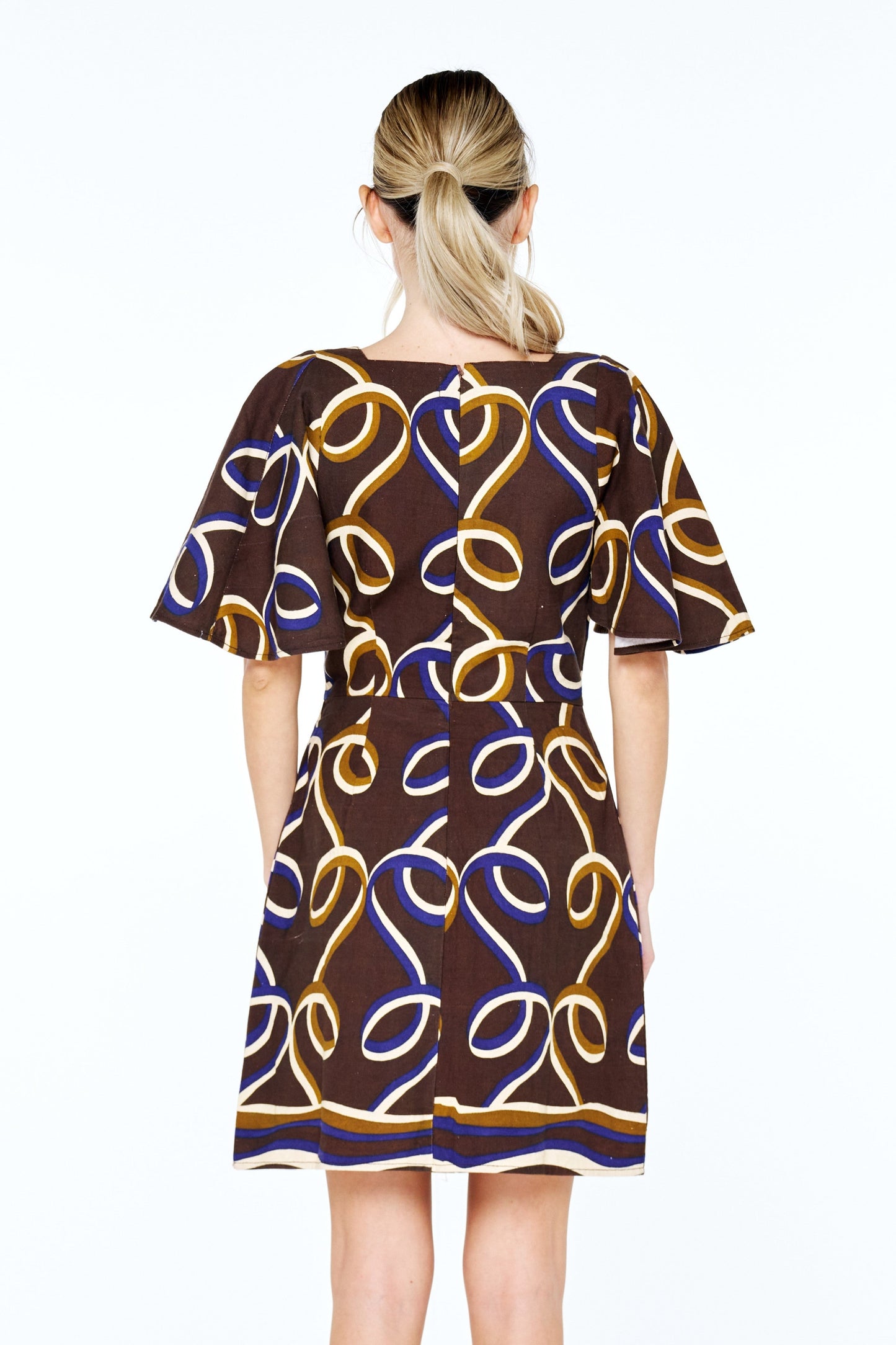 A woman viewed from the back wearing a short-sleeve A-line dress with a brown base and an abstract wavy line pattern in yellow, blue, and white. She is wearing black strappy sandals against a plain white studio background.
