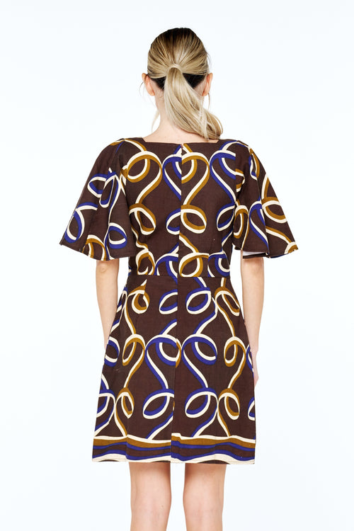 Load image 1 in gallery view. A woman viewed from the back wearing a short-sleeve A-line dress with a brown base and an abstract wavy line pattern in yellow, blue, and white. She is wearing black strappy sandals against a plain white studio background.