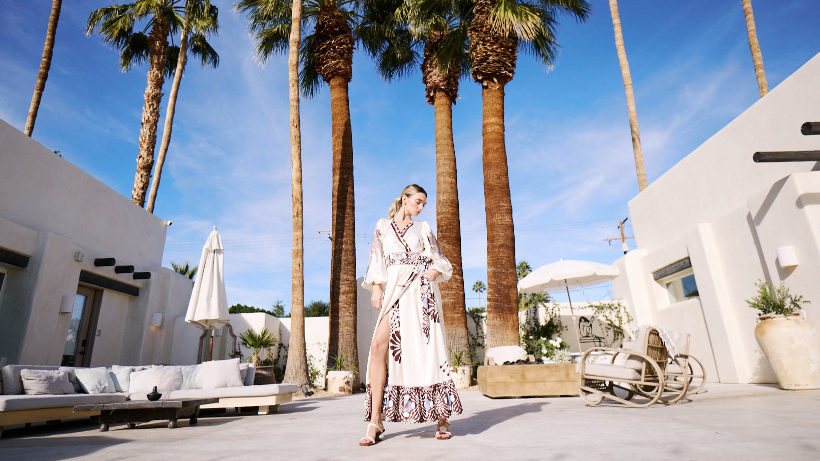 Woman in a white dress standing in a tropical outdoor setting with palm trees and white buildings.