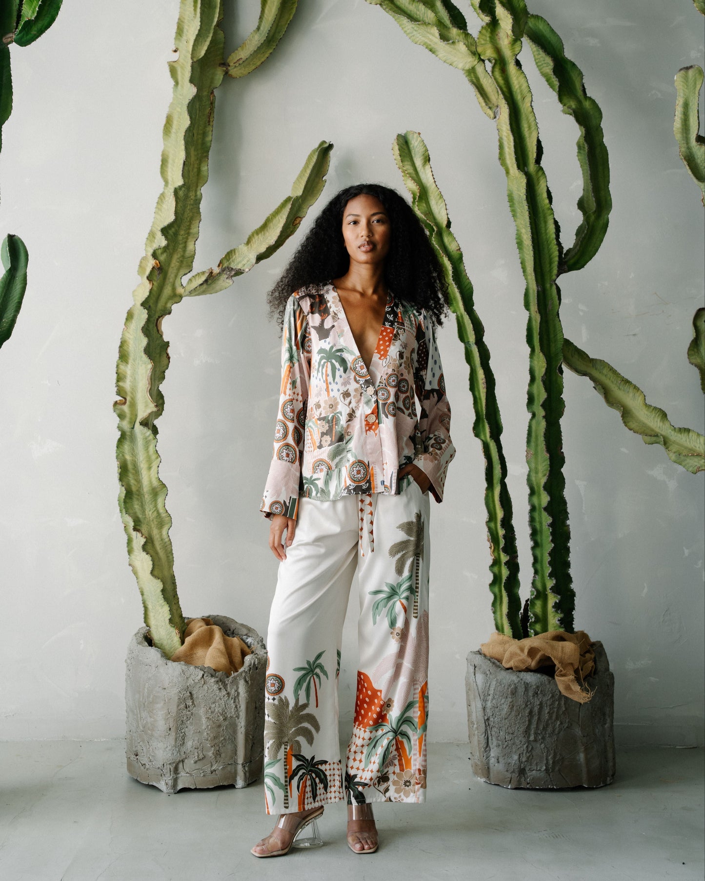Woman in a floral outfit standing between two tall cacti against a white wall.