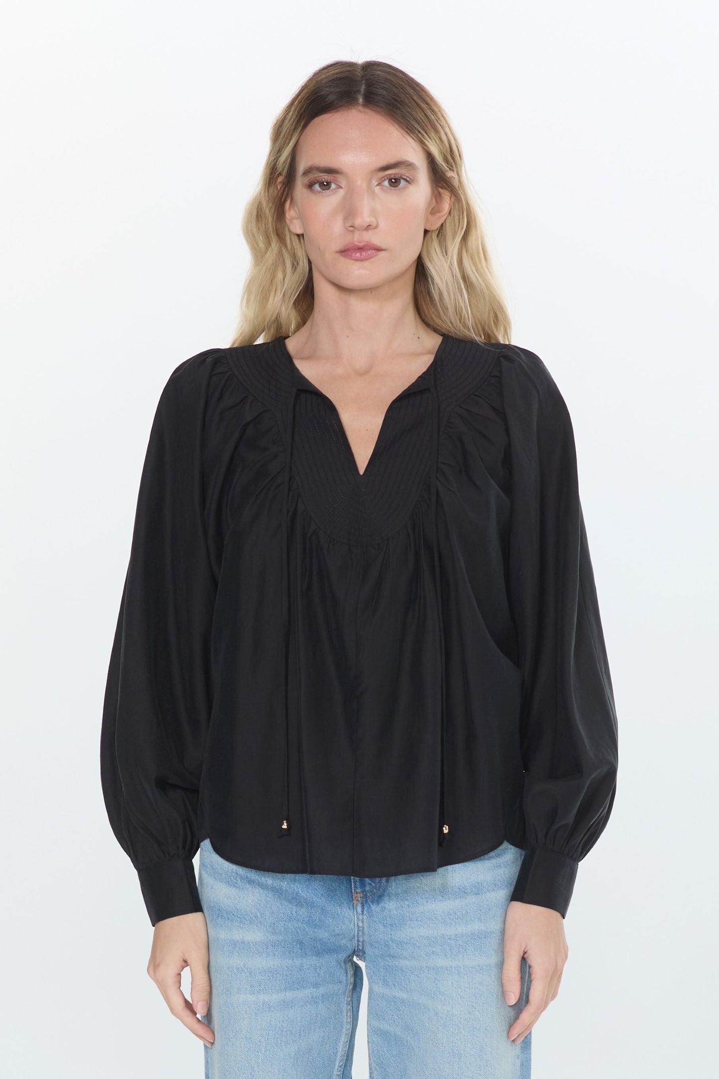 A woman stands against a plain white studio background wearing a black long-sleeve blouse with a gathered V-neck and light-wash blue jeans.