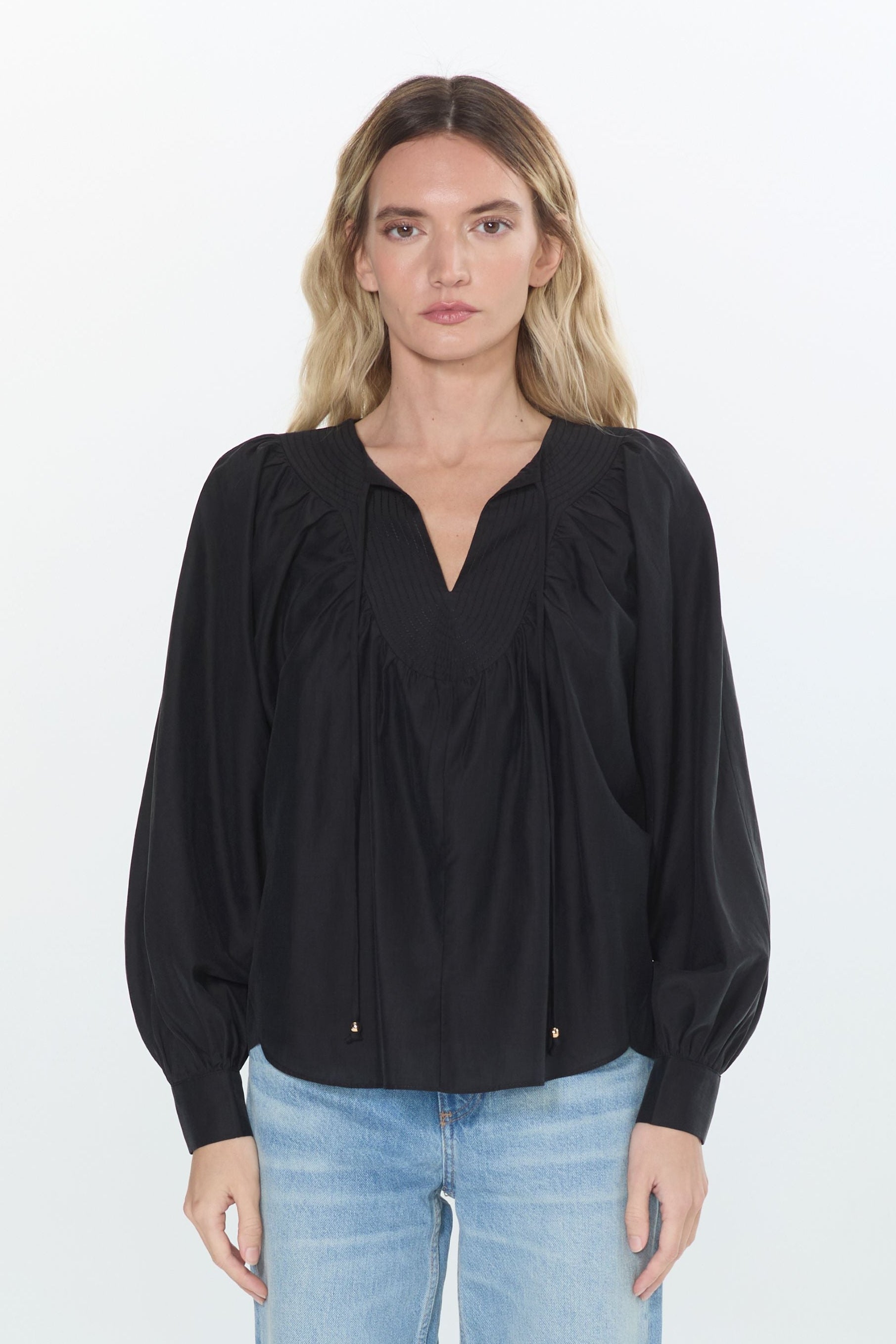 A woman stands against a plain white studio background wearing a black long-sleeve blouse with a gathered V-neck and light-wash blue jeans.