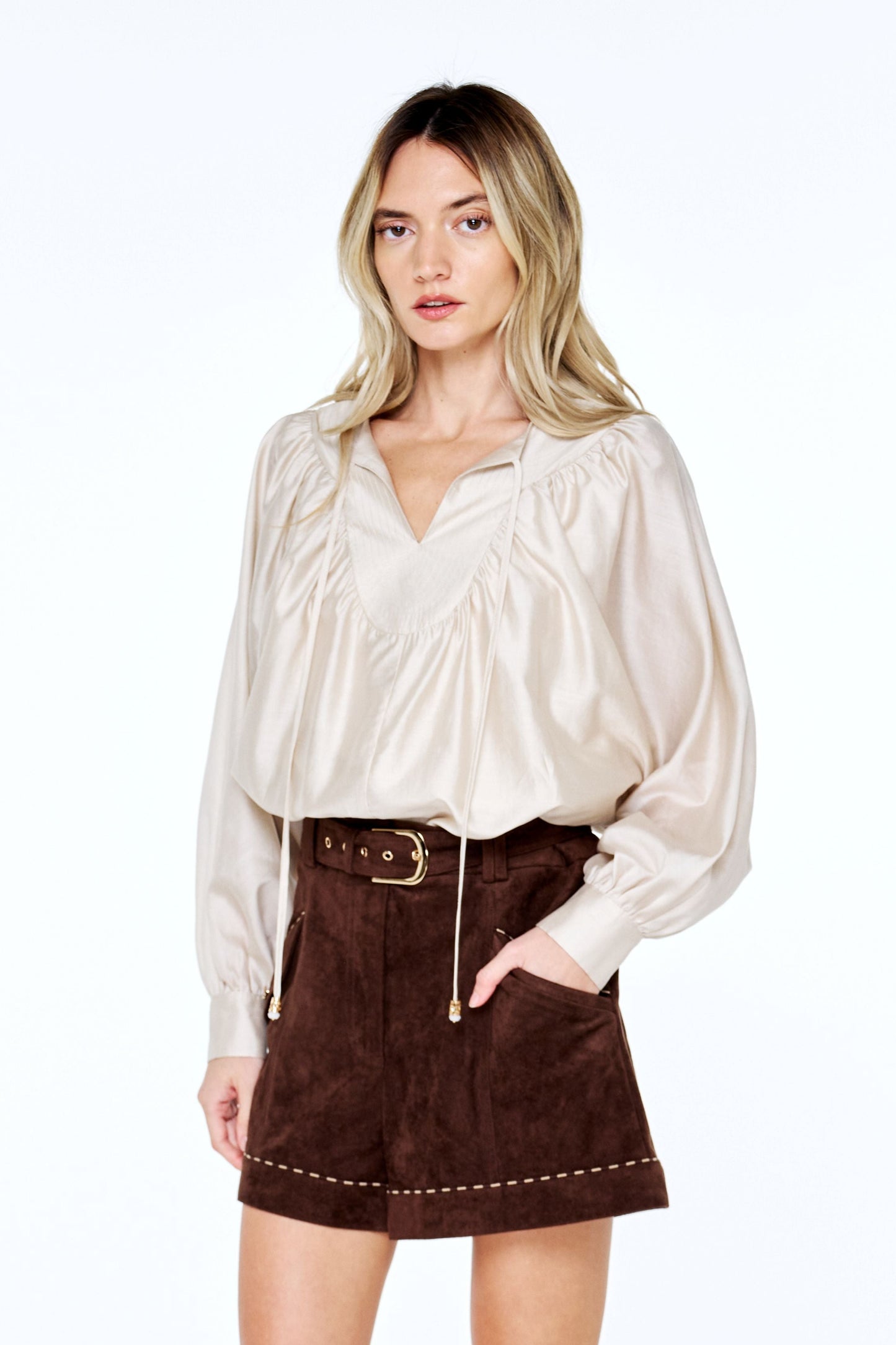 A woman modeling a cream satin blouse and dark brown suede shorts with a matching belt, standing against a plain white studio background.