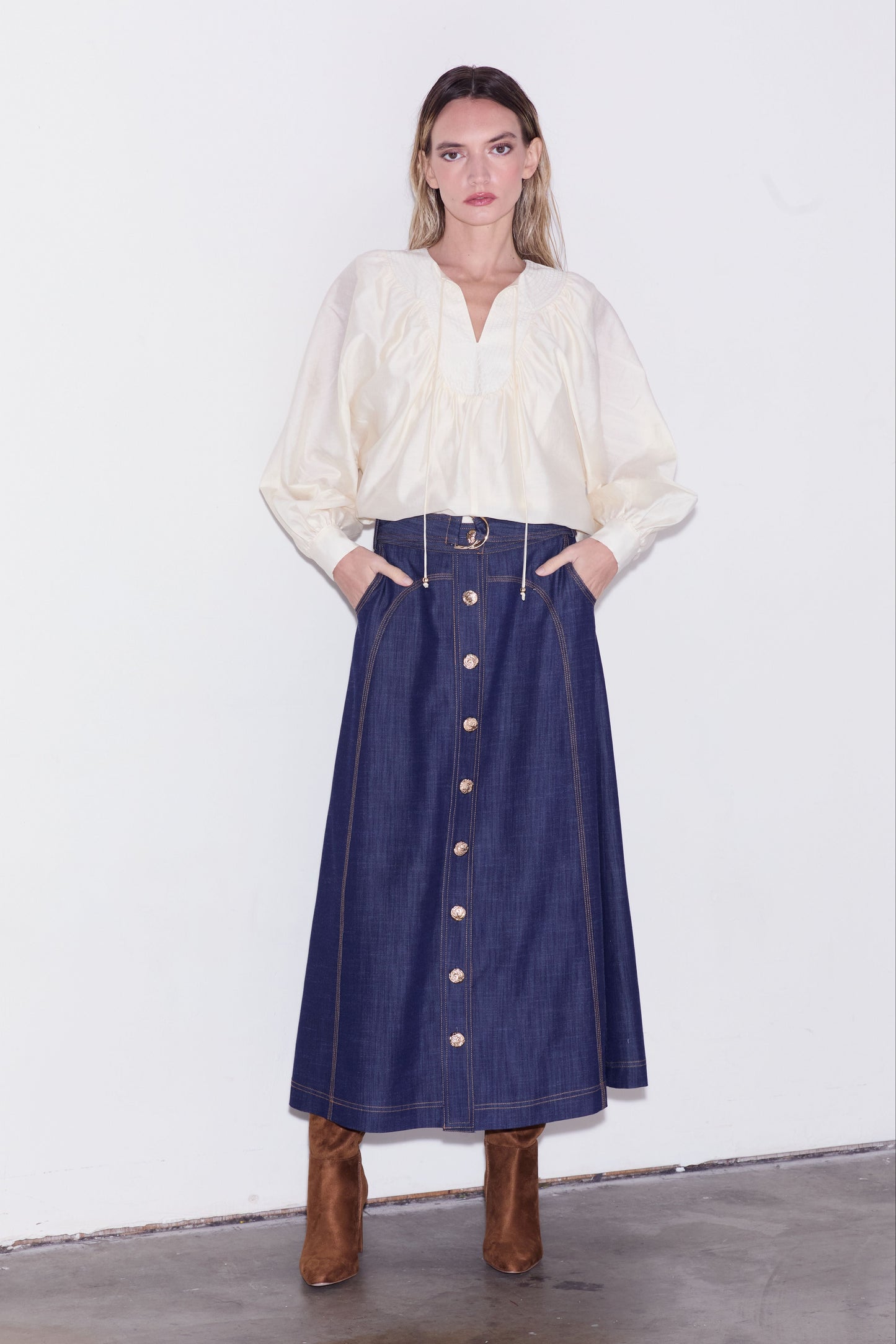 A model stands against a white wall wearing a cream-colored long-sleeve blouse and a dark blue denim midi skirt with gold buttons, paired with brown boots.