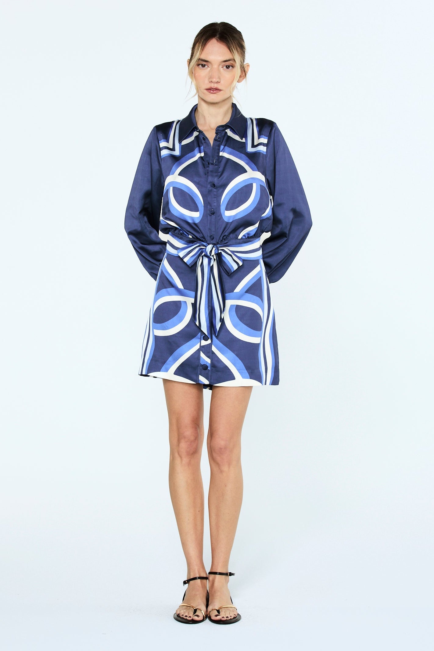 A model stands against a plain white studio background wearing a short navy satin-like shirt dress with a bold white and light blue geometric pattern and a self-tie belt.