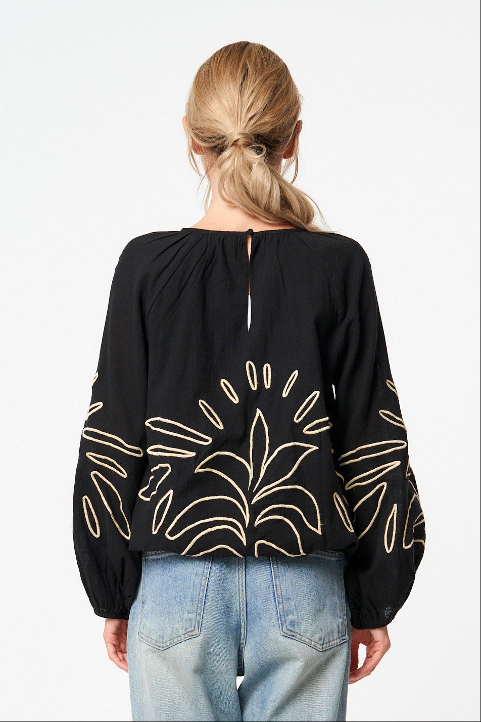 A woman viewed from the back wearing a black long-sleeve blouse with gold leaf embroidery on the sleeves and light-wash blue jeans, standing against a plain white studio background.