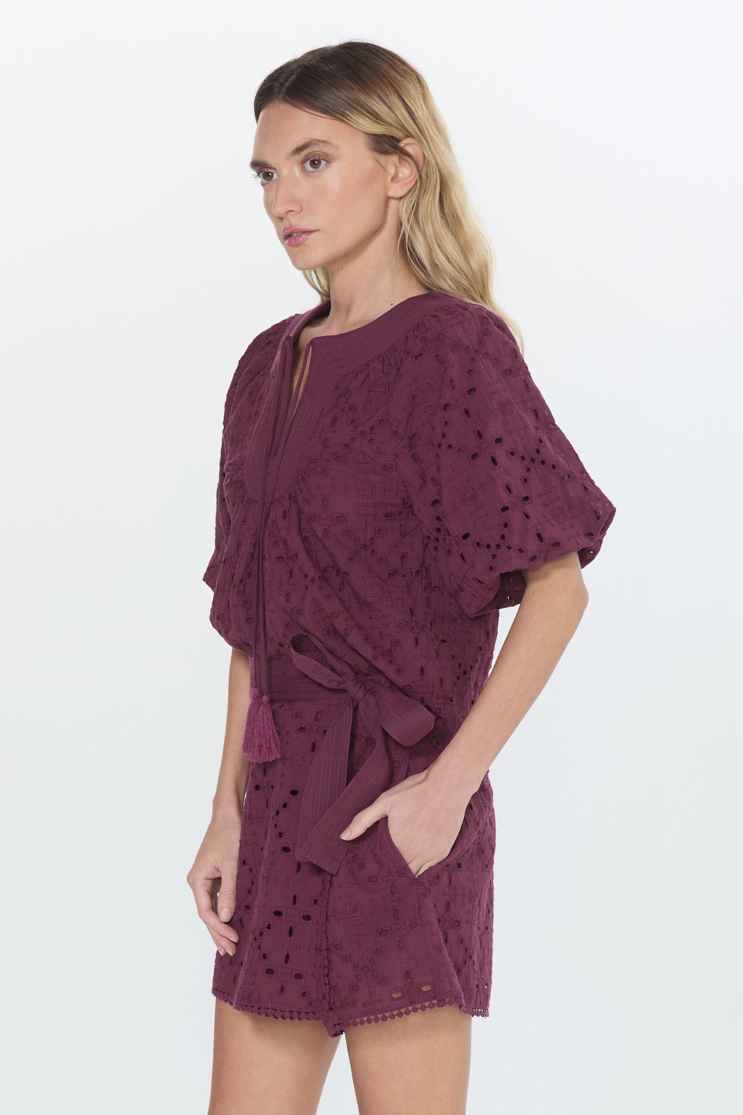 A woman modeling a short, loose-fitting eyelet dress in deep burgundy with a self-tie waist belt, paired with black flat sandals, against a plain white studio background.